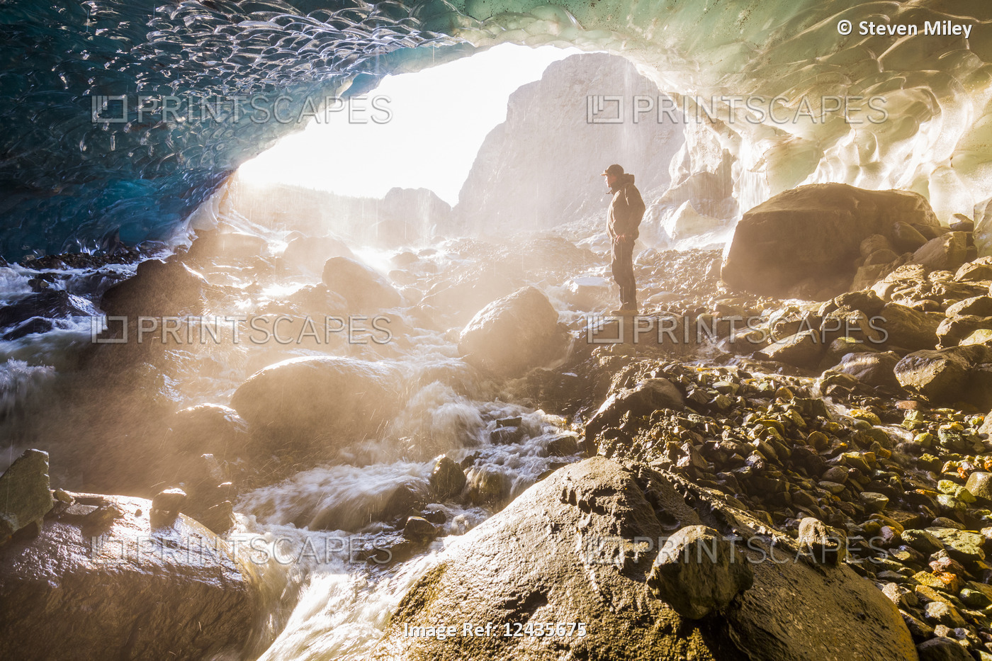 A man stands at the entrance of an ice cave bathed in warm sunlight and mist, ...
