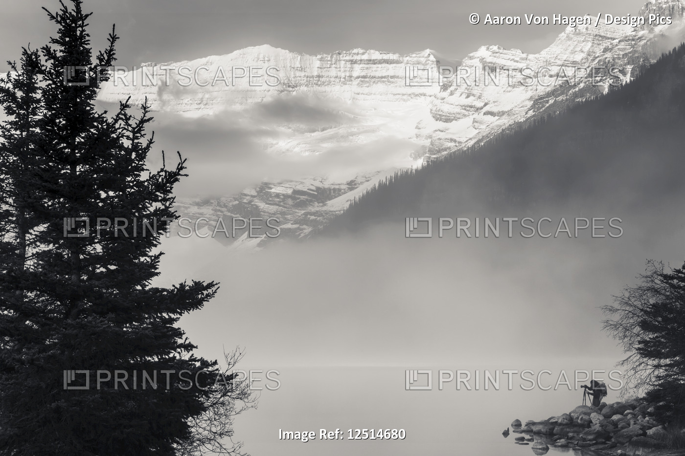 Silhouette of a photographer on the shore of Lake Louise with fog rising from ...
